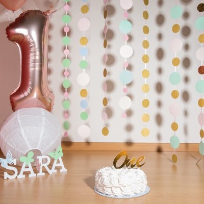 Decoração de festa com balão número 1 rosa, nome SARA em madeira branca, bolo branco com topo dourado 'One' e cortinas coloridas