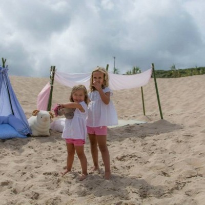 Duas meninas em roupa clara na areia junto a tendas de tecido azul e branco com almofadas e urso de pelúcia