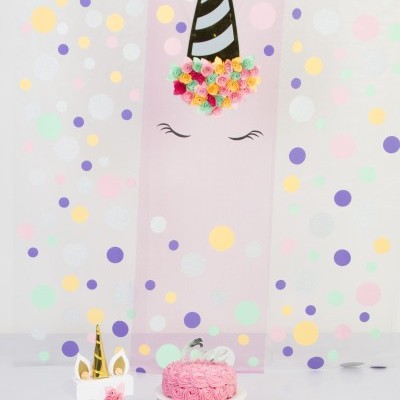 Decoração com tema unicórnio rosa, chifres dourados, vela número 7, e bolo cor-de-rosa em suporte branco.