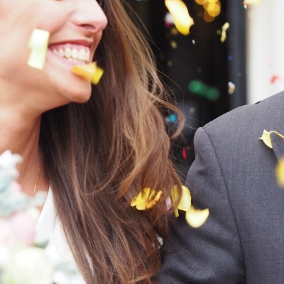 Mulher sorridente com cabelo castanho e camisa branca ao lado de homem com fato cinzento com pétalas coloridas ao redor