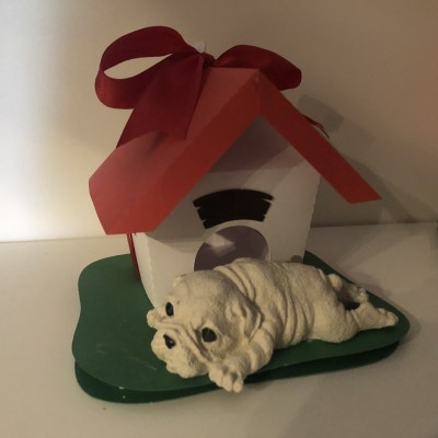 Escultura de cachorro deitado à frente de casinha com telhado vermelho e laço