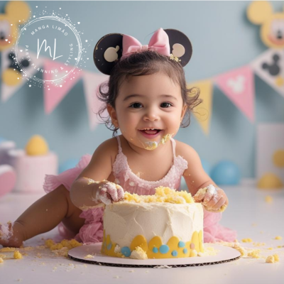 Bebé com faixa de orelhas de rato e vestido rosa a comer bolo branco decorado com amarelo e azul