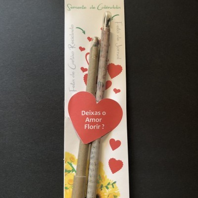 Pacote com dois lápis de cartão reciclado com sementes de calêndula e embalagem branca com corações vermelhos e texto promocional.