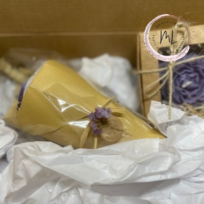 Embrulho de presente com papel amarelo, fita amarela e flor roxa seca num ambiente com papel de seda branco e caixa de embalagem marrom