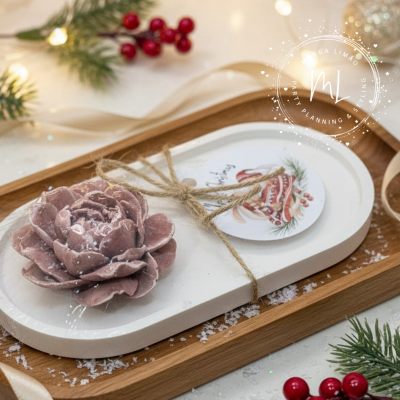 Ornamento decorativo natalício com flor seca roxa numa base oval branca com etiqueta de sinos e ramos verdes, sobre bandeja de madeira e decoração festiva.