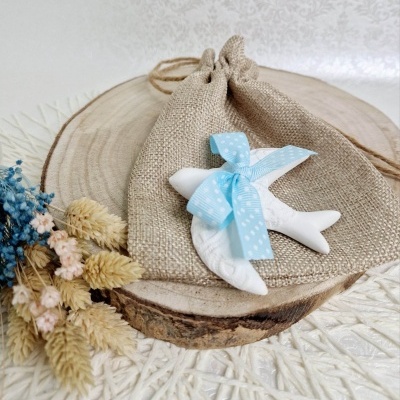 Saco de tecido com figura de peixe cerâmica e laço azul sobre placa de madeira redonda com ramo de flores secas