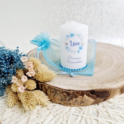 Vela branca decorativa para primeira comunhão sobre disco de madeira com flores secas e saco de organza azul