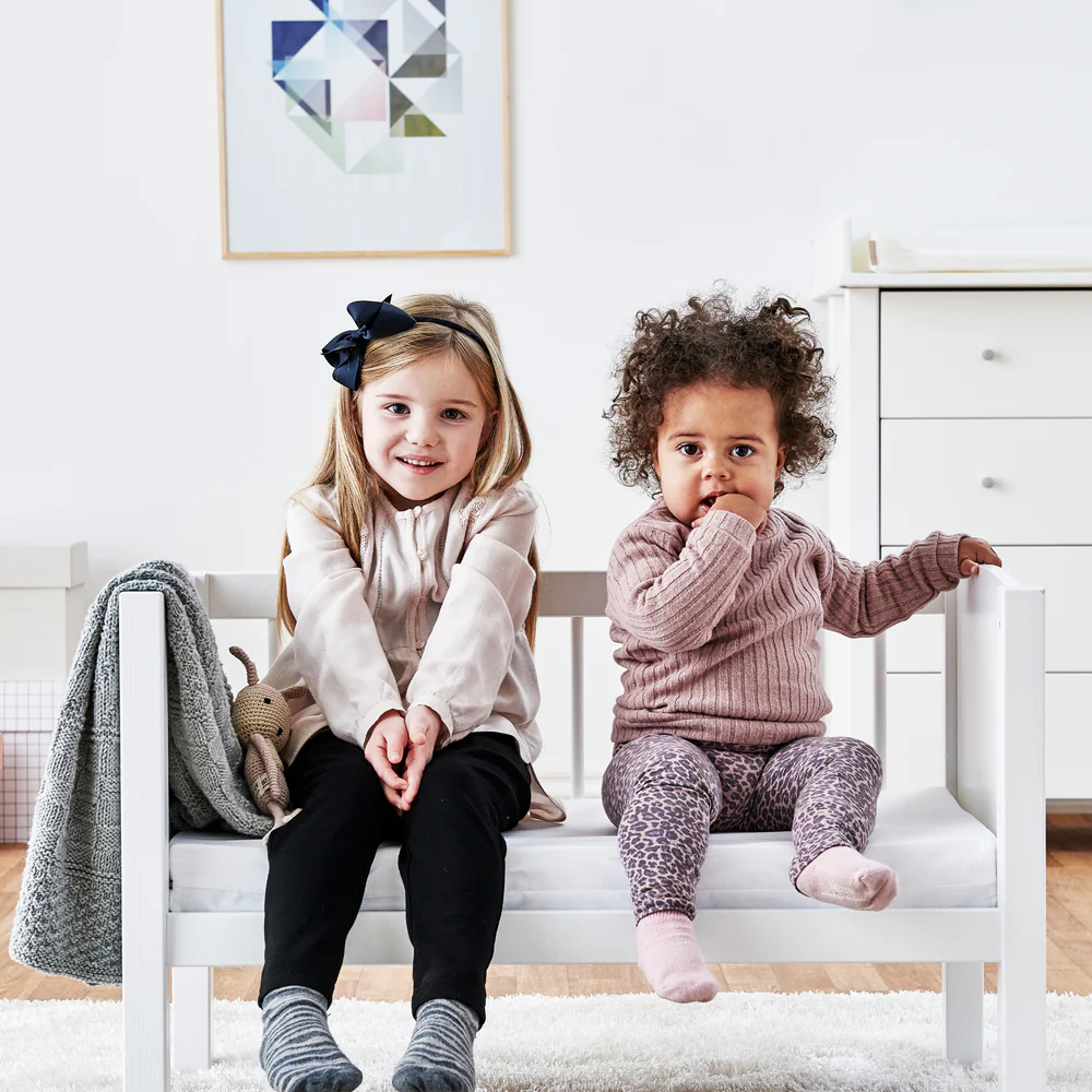 Colchão de Espuma para Berço e Cama de Grades Duas crianças sentadas num sofá branco de madeira, uma com cabeleira loira e outra com cabelo encaracolado.