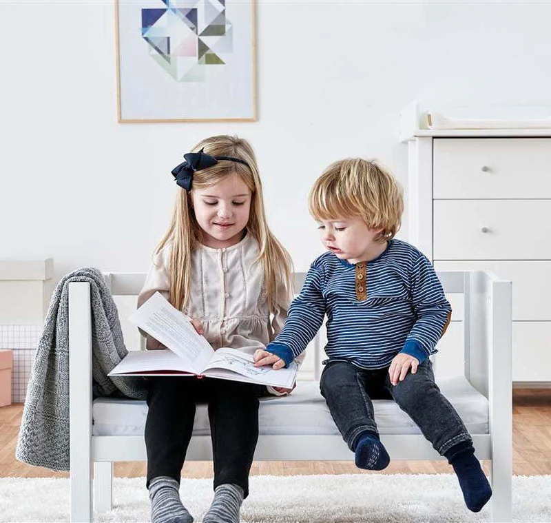 Colchão de Espuma para Berço e Cama de Grades Cama infantil branca de madeira com duas crianças sentadas lendo em quarto minimalista branco.