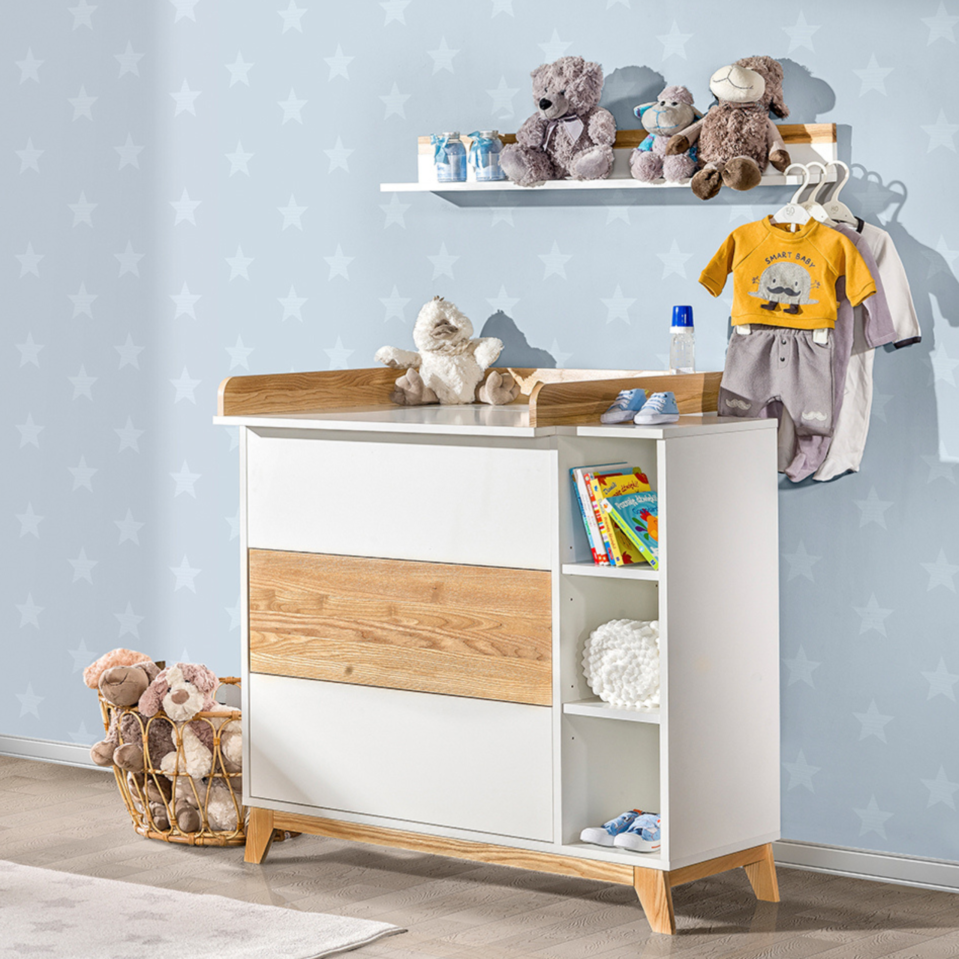 Cómoda com Trocador e Gavetas NORDIK Cômoda infantil branca com detalhes em madeira, peluches, livros e roupa de bebé num quarto com parede azul de estrelas.