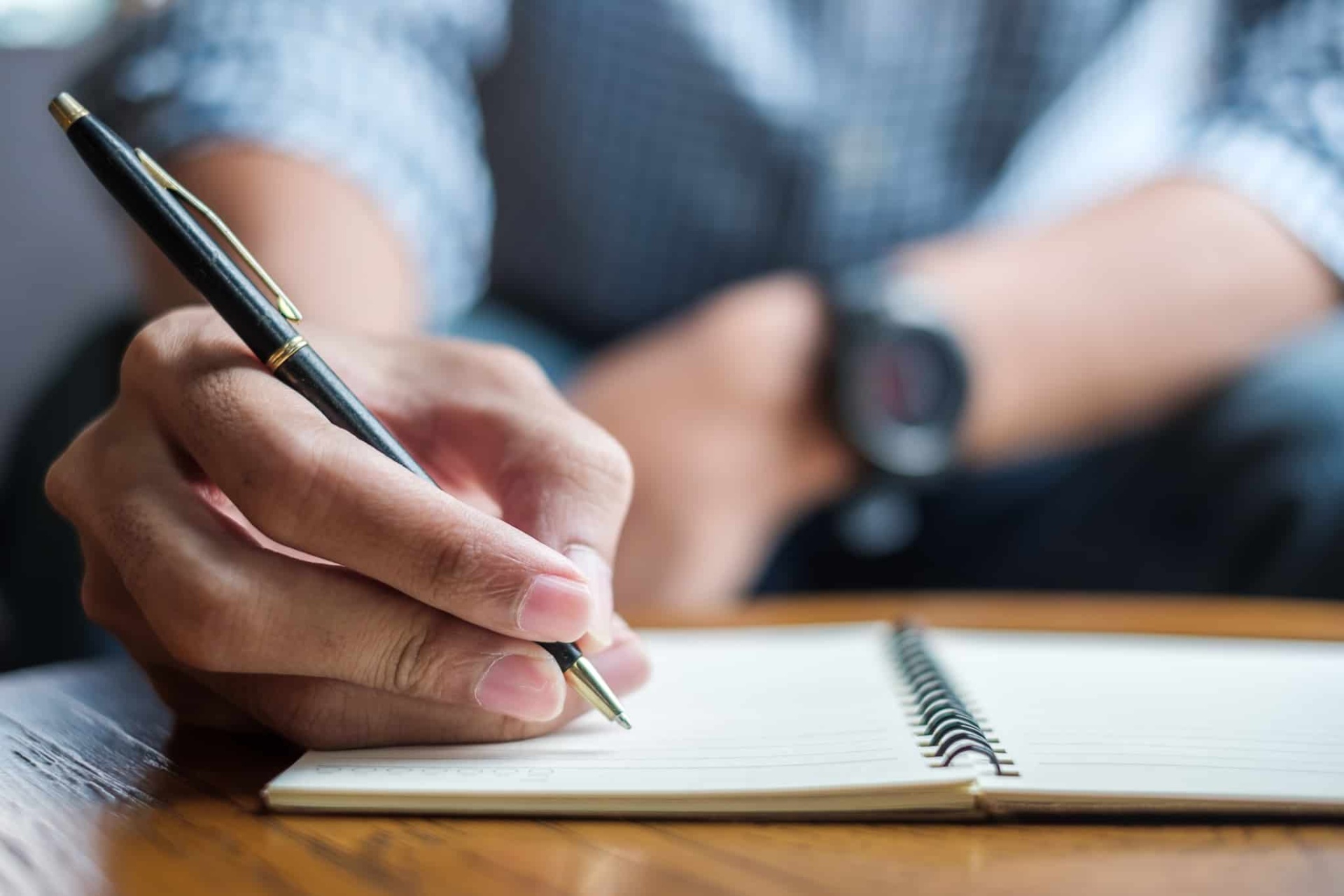 Pessoa a escrever num caderno de espiral com caneta preta e detalhes dourados sobre mesa de madeira