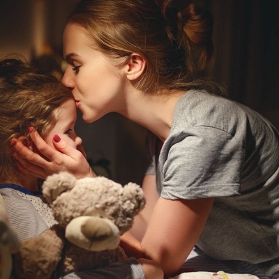 Mulher a beijar criança na testa, criança segura urso de peluche na cama