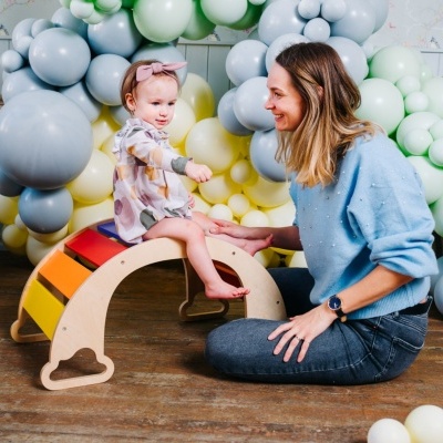 Criança sentada num baloiço de madeira colorido com balões ao fundo e adulta ao lado