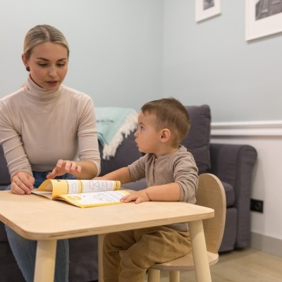 Mesa de madeira clara com livro aberto e duas cadeiras infantis numa sala com sofá cinza
