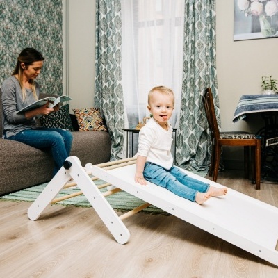 Tobogã infantil branco em sala de estar com criança a brincar e mulher sentada no sofá.
