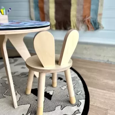 Cadeira infantil de madeira clara com encosto em forma de orelhas de coelho junto a mesa e tapete redondo preto e cinza