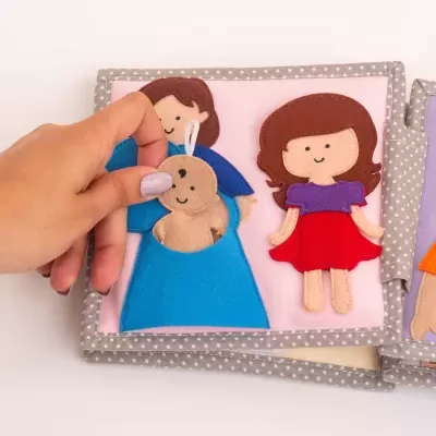 Livro têxtil infantil com bonecas de feltro colorido e padrão de bolinhas cinza.