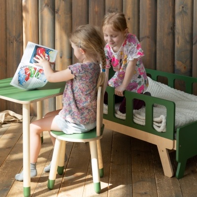 Cama infantil verde com colcha branca, mesa e cadeira de madeira clara, duas crianças brincando
