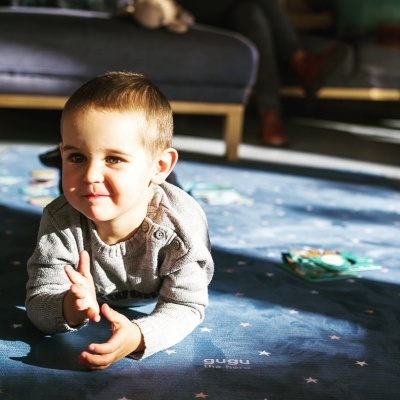 Criança deitada num tapete infantil azul escuro com estrelas brancas e texto 'gugu The Rug'