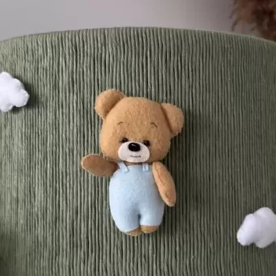 Ursinho de peluche com macacão azul claro num fundo verde com nuvens brancas