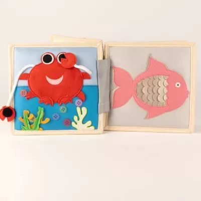 Livro de tecido com caranguejo vermelho e peixe rosa em páginas opostas