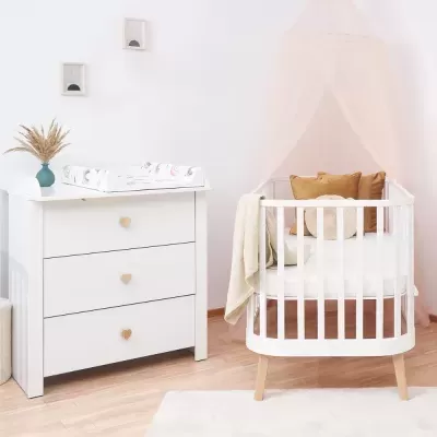 Móvel branco com três gavetas e cómoda com trocador ao lado de berço branco com almofadas e dossel rosa