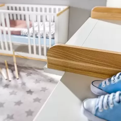 Berço branco com detalhes em madeira, tapete cinza com estrelas e sapatos azuis de bebé em superfície branca.