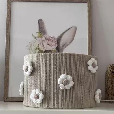 Abajur bege com flores decorativas em corda, em prateleira branca, com quadro de orelhas de coelho atrás
