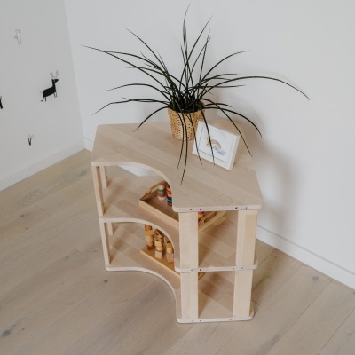 Mesa triangular em madeira clara com planta em vaso amarelo sobre ela num chão de madeira clara junto a parede branca com autocolantes.