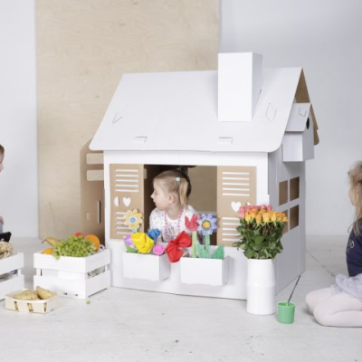 Casa de brincar de cartão branco com três crianças e caixas com brinquedos e plantas coloridas num ambiente interior