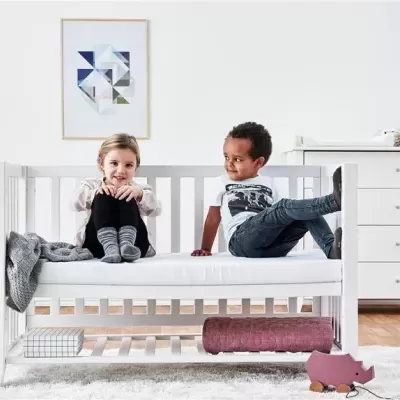 Berço branco em madeira com colchão branco e duas crianças sentadas