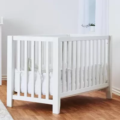 Berço branco de madeira com colchão branco e fronhas estampadas, em ambiente interior com chão de madeira.