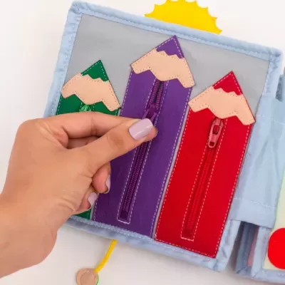 Livro educativo de tecido com lápis coloridos e fechos de correr sendo aberto por uma mão com unhas lilás.