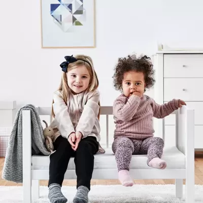 Duas crianças sentadas num sofá branco de madeira, uma com cabeleira loira e outra com cabelo encaracolado.