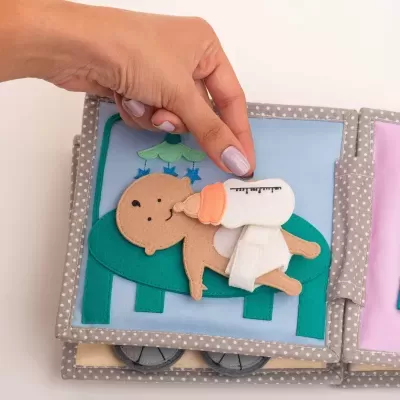 Livro têxtil infantil com figura de bebê a ser alimentado com biberão.