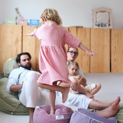 Criança em vestido rosa está em almofada rosa, com almofada lilás ao lado e adultos no sofá ao fundo