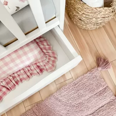 Berço branco com almofada rosa aos quadrados na gaveta, tapete rosa peludo e chão de madeira clara.