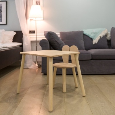 Mesa e cadeira infantis de madeira clara junto a sofá cinzento e cama num quarto acolhedor.