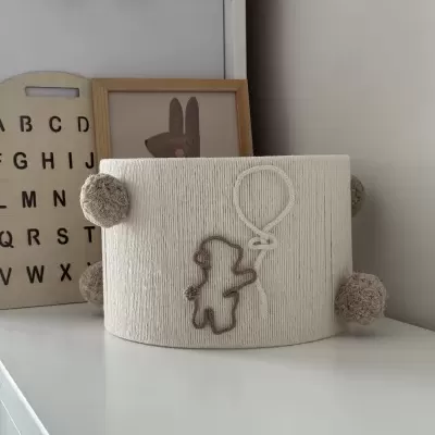 Artigo decorativo em tecido bege com pompons castanhos e ursinho com balão.