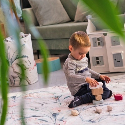 Criança a brincar com blocos de madeira num tapete floral em sala com sofá e casinha de brinquedo