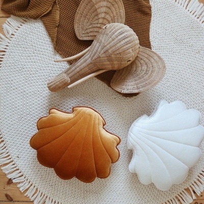 Tapete redondo branco com franjas, almofadas em forma de concha bege e branca, manta castanha e ventilador de vime