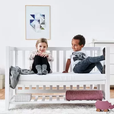 Berço branco de madeira com crianças sentadas e cobertor cinza na lateral