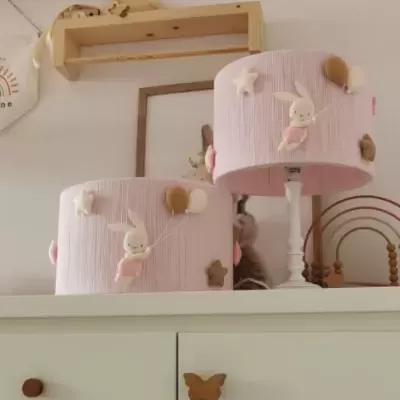 Duas lâmpadas de mesa com abajur rosa decorado com coelhos e balões sobre cómoda branca com puxadores de borboleta