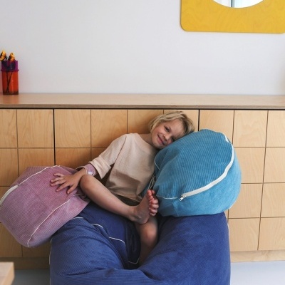 Jovem sentado em almofadas grandes de tecido riscado azul, rosa e azul claro junto a cómoda de madeira clara