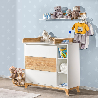 Cômoda infantil branca com detalhes em madeira, peluches, livros e roupa de bebé num quarto com parede azul de estrelas.