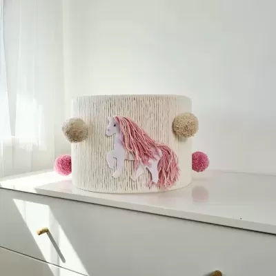 Caixa redonda decorativa com cavalo rosa e pompons sobre cómoda branca