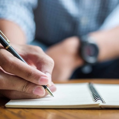 Pessoa a escrever num caderno de espiral com caneta preta e detalhes dourados sobre mesa de madeira
