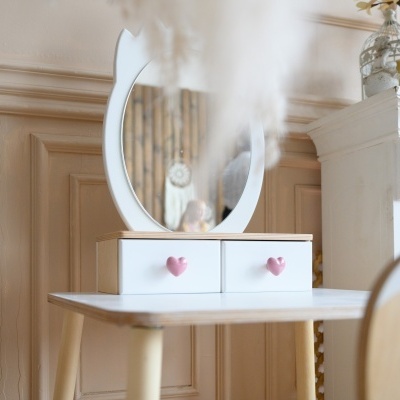 Móvel branco com duas gavetas e espelho redondo com puxadores em forma de coração cor-de-rosa.