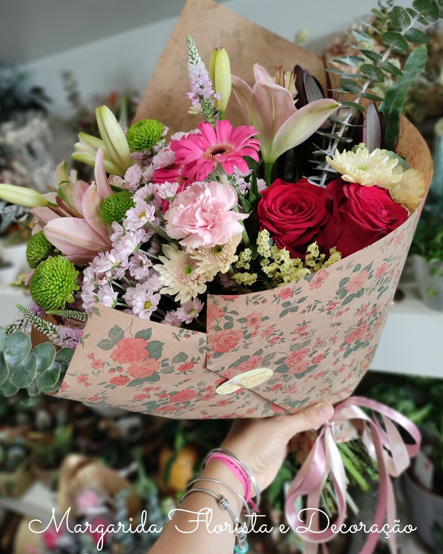 Buquê colorido de flores variadas com envelope floral rosa segurado por mão com pulseiras