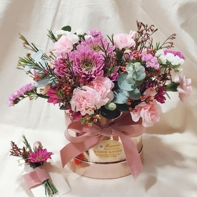 Arranjo floral colorido em caixa redonda dourada e mini buquê ao lado sobre tecido branco